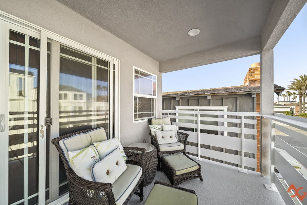 A covered outdoor balcony with wicker seating, including two armchairs and an ottoman, faces a white slatted railing and offers a partial view of neighboring rooftops and palm trees. Sliding glass doors and a window provide access to the interior.