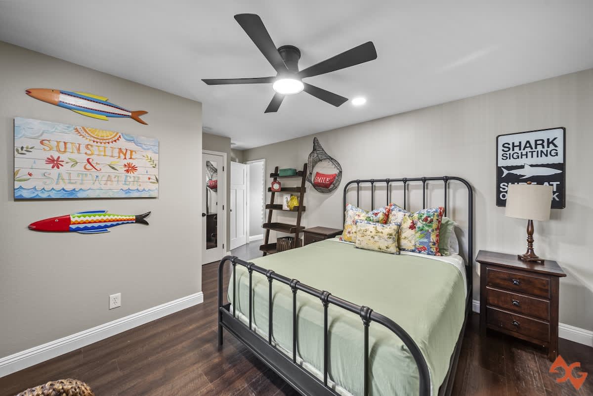 A bedroom with a black metal bed frame, light green bedding, and vibrant floral pillows, complemented by a black ceiling fan and coastal-themed wall decor including fish and a "Shark Sighting" sign.