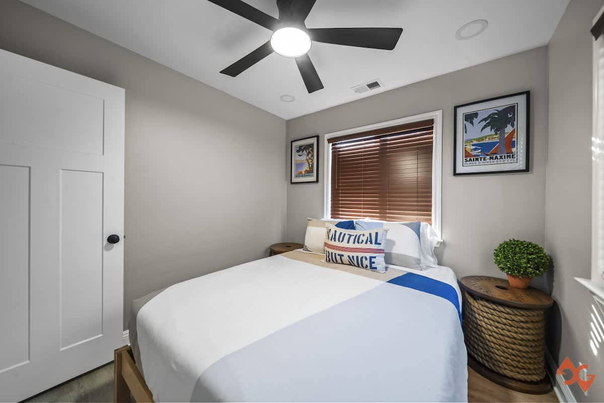 A view of bedroom-1 showing a queen bed with blue and white bedding, a rope-wrapped side table with a plant, and a white door. Two windows with dark wood blinds and framed beach-themed art are visible.