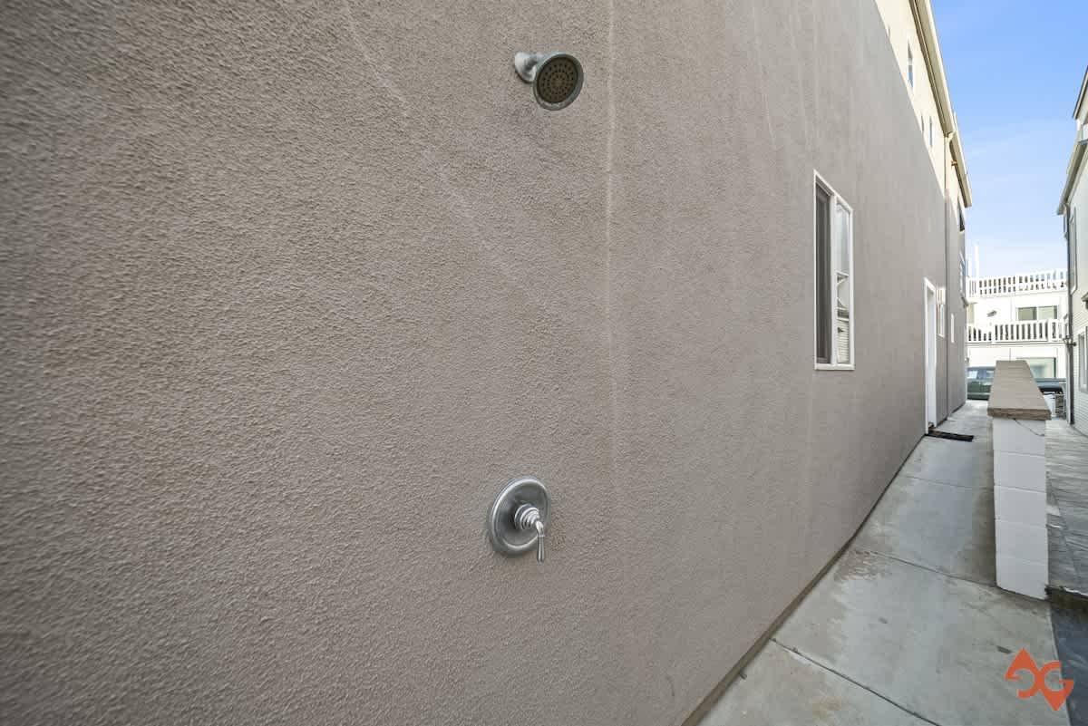 A textured stucco wall features an outdoor shower with a modern chrome shower head and a single handle faucet, set above a clean concrete path.