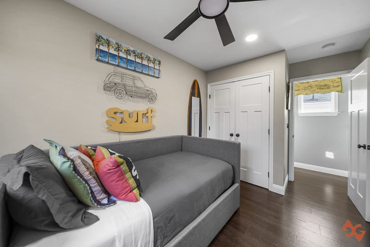 A surf-themed bedroom with a gray daybed, dark wood floors, and a ceiling fan. The walls are decorated with a surfboard, a 'Surf' sign, and a car illustration, creating a playful coastal atmosphere.