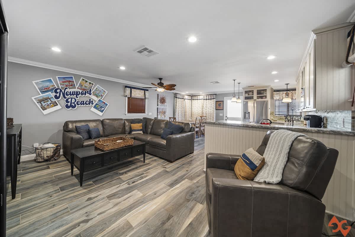 Spacious living room featuring a large gray leather sectional sofa, a dark coffee table, and an additional armchair, all on light wood-look flooring with coastal-themed wall art.