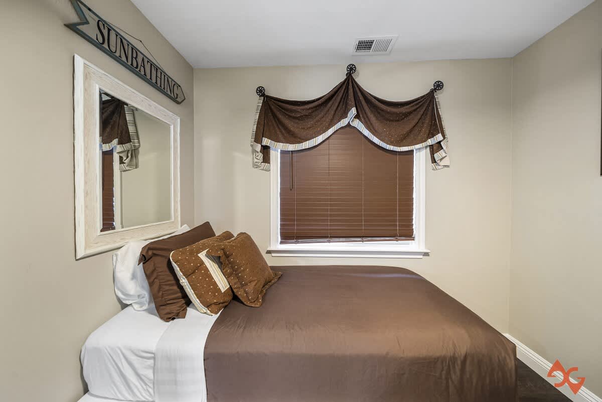 A bedroom with a bed featuring brown bedding and a white storage base, positioned under a window with brown blinds and a decorative valance. Dark wood floors and a white framed mirror are also visible.