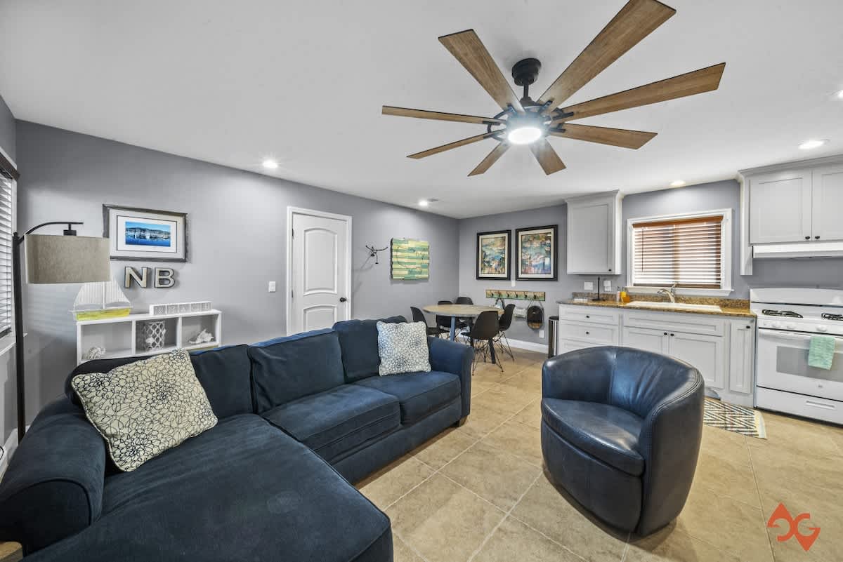Open-concept living and dining area with a blue sectional sofa, a round dining table, and a white kitchen, all on light tile flooring, featuring a large multi-blade ceiling fan.