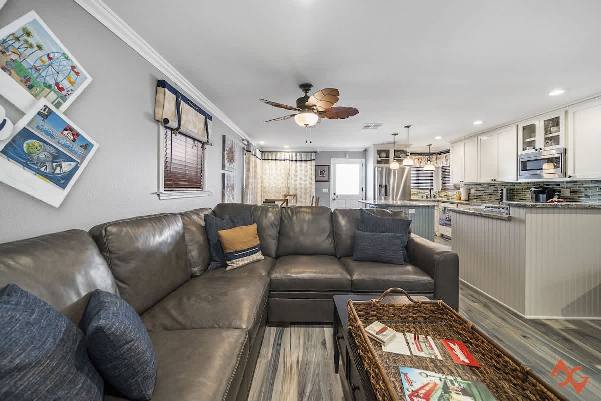 Open concept living space featuring a large gray leather sectional sofa with blue throw pillows, facing a kitchen with light wood-look flooring and a ceiling fan.