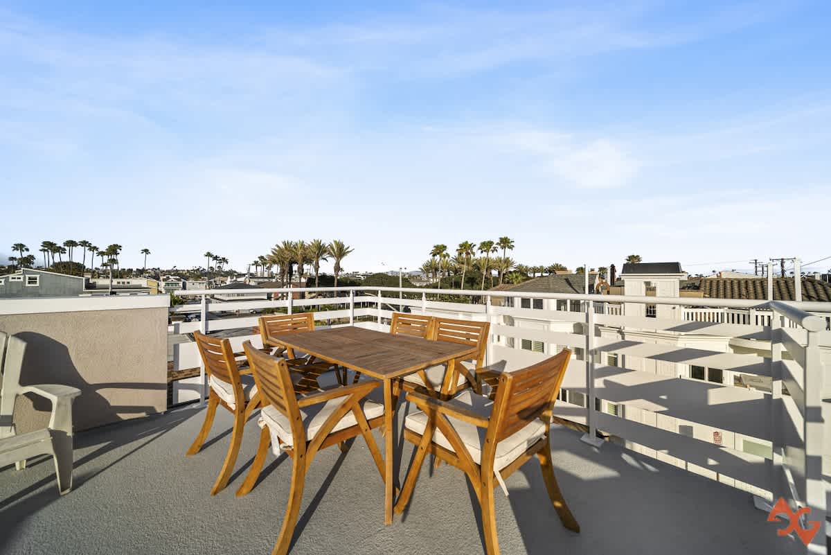 A wooden outdoor dining set with six chairs and cushions is positioned on the rooftop deck, offering an open view of the surrounding coastal neighborhood with numerous palm trees and residential buildings.