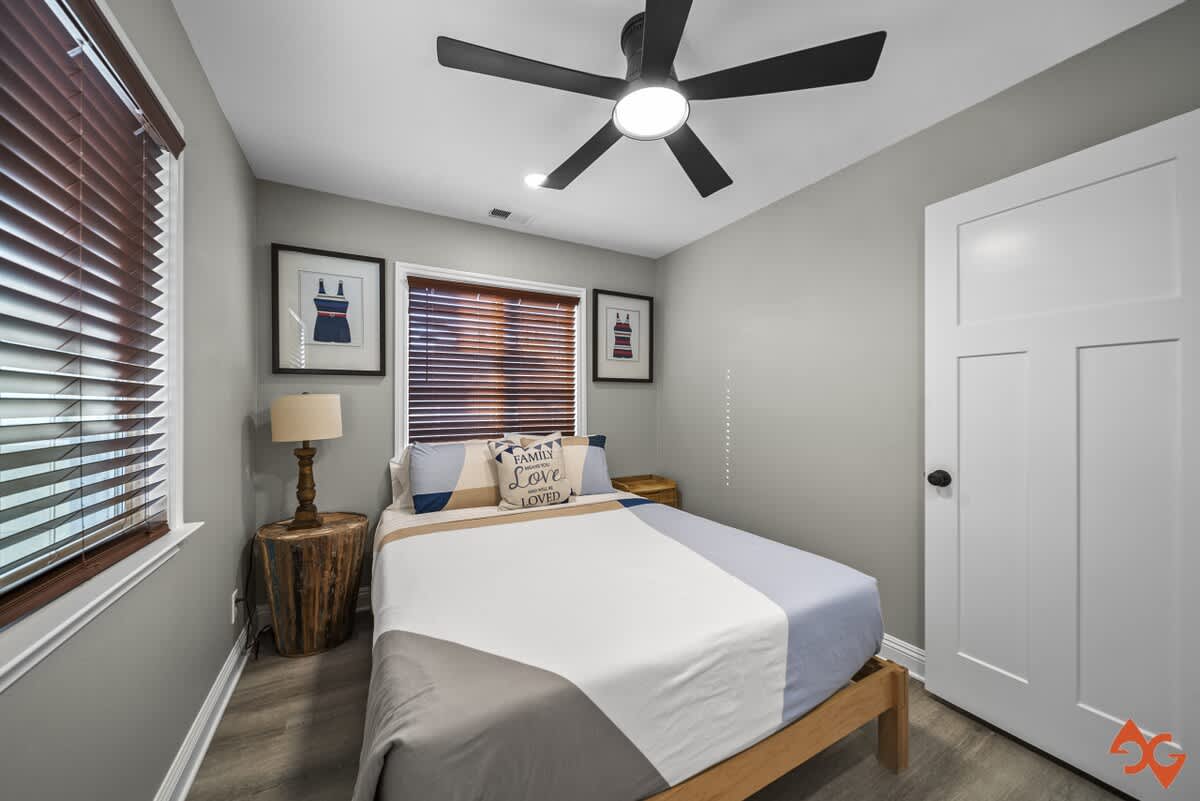 A wider view of bedroom-2, showcasing a queen bed with blue and white bedding, a wooden nightstand with a lamp, and two windows with dark wood blinds. Framed swimsuit art and a white door are visible.