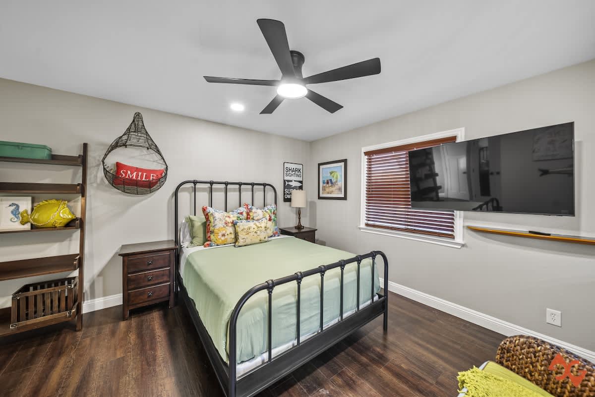 A bright bedroom with a black metal bed frame, a dark wood nightstand with a lamp, a wall-mounted TV, and a window with wooden blinds, all under a black ceiling fan.