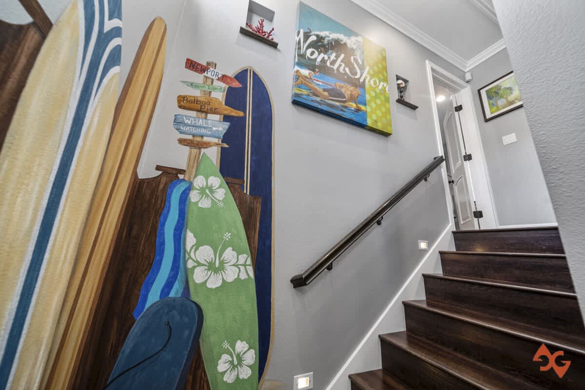 A vibrant staircase area featuring dark wood steps with recessed lighting and a dark handrail. The wall is adorned with a colorful surfboard mural and beach-themed artwork, creating a lively coastal ambiance.