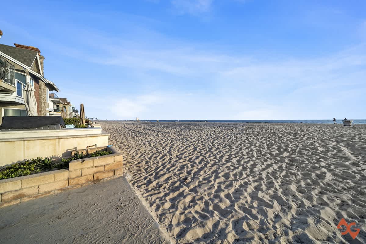 A wide expanse of sandy beach stretches towards the ocean under a bright blue sky, with a glimpse of a building on the left and distant structures along the coastline.