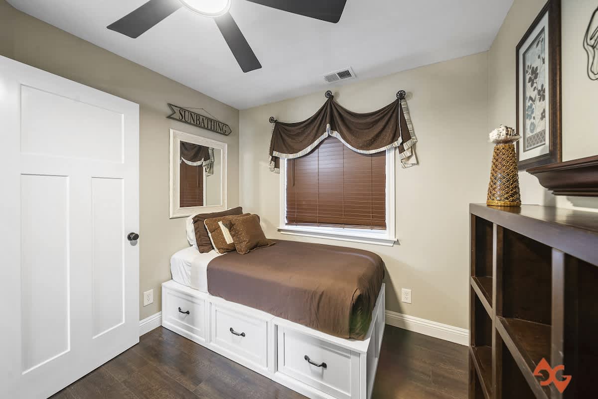 A cozy bedroom featuring a bed with brown bedding and a white storage base, dark wood floors, and a ceiling fan. A white framed mirror hangs on the wall next to a white door, and the window has brown blinds.