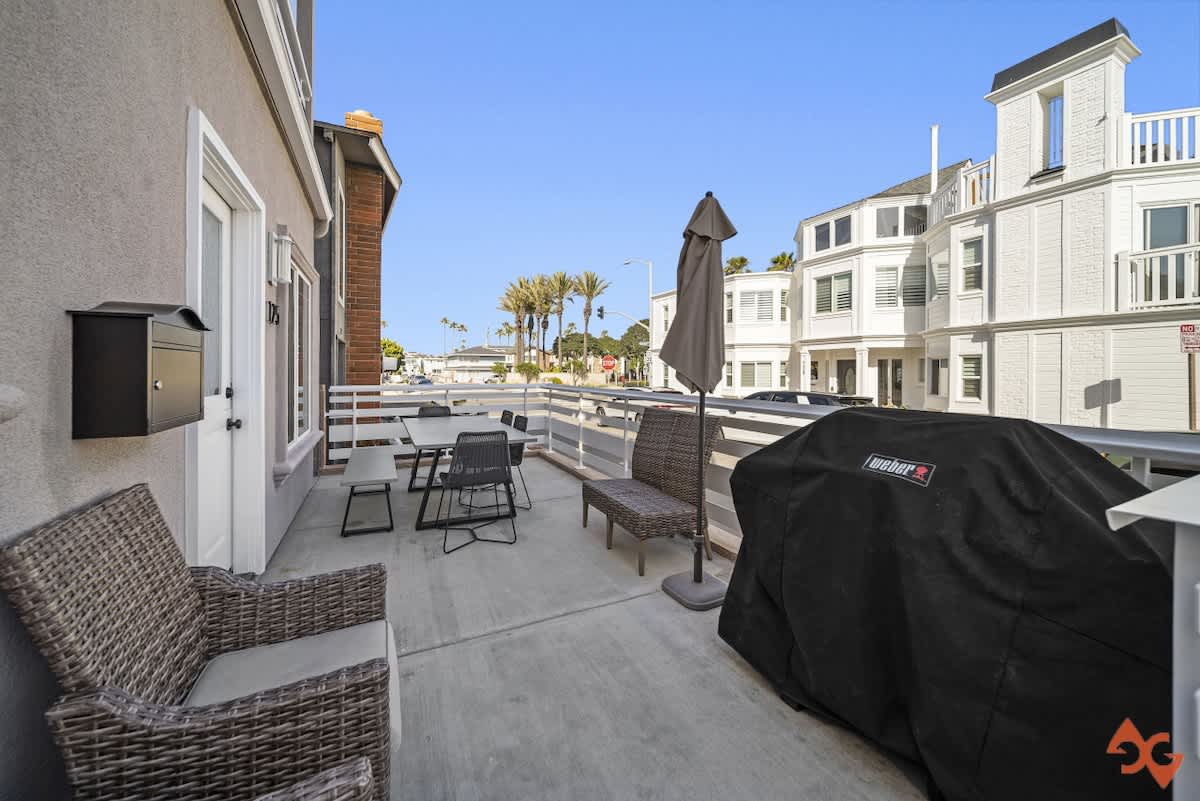 A front patio area with a white slatted railing features a covered BBQ grill, an outdoor dining table with chairs, and wicker seating, offering views of the street and neighboring houses.