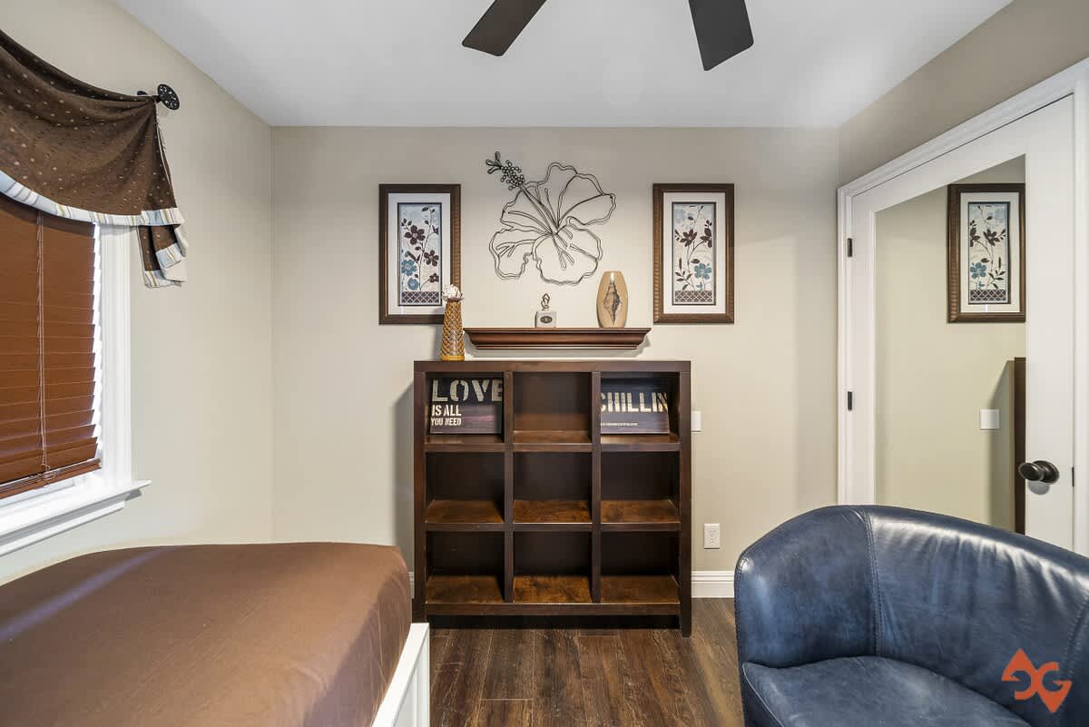 A bedroom with dark wood floors and light-colored walls, featuring a dark wood bookshelf filled with decorative items. Artwork and a metal floral wall decor are displayed above the bookshelf, adding a touch of elegance.