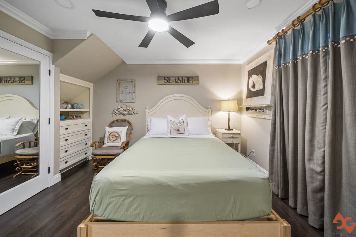 A cozy bedroom with a light green bedspread on a white bed frame, dark wood floors, and a ceiling fan. A white dresser with open shelving and a chair are visible on the left, and a nightstand with a lamp on the right.