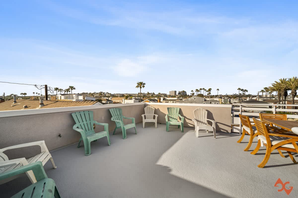 A wide shot of the rooftop deck showcasing a row of light green and white Adirondack chairs along one wall and a wooden outdoor dining set, with a backdrop of residential rooftops and numerous palm trees.