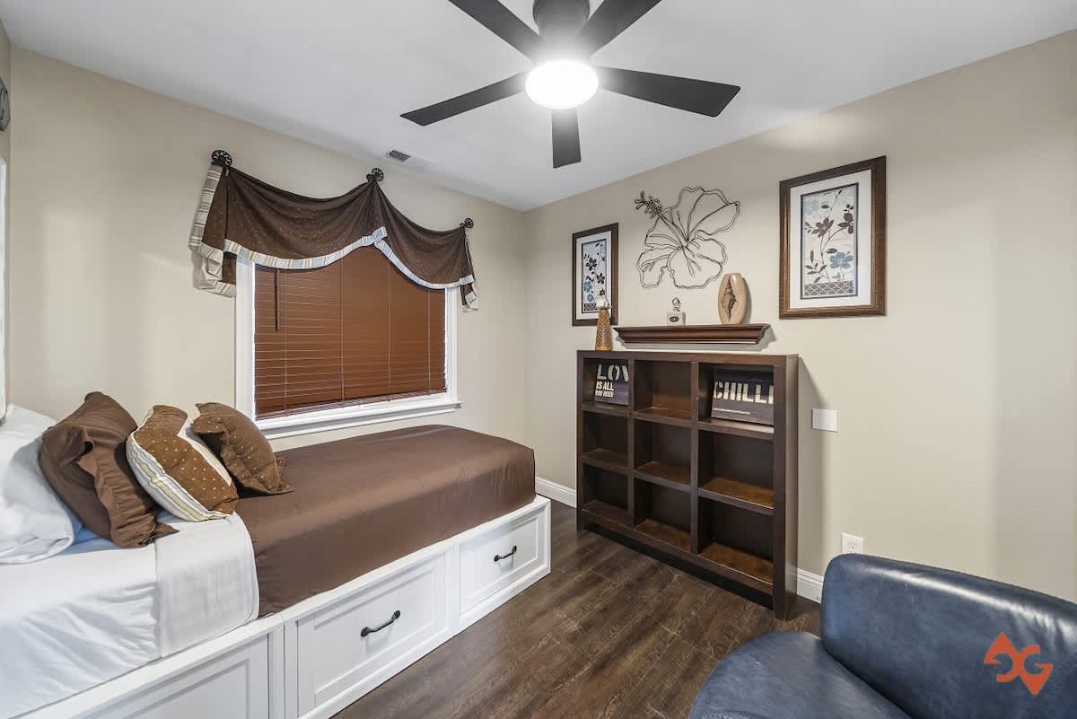 A bedroom with dark wood floors, a bed featuring brown bedding and a white storage base, and a dark wood bookshelf. A dark blue armchair is partially visible, and the window has brown blinds and a decorative valance.