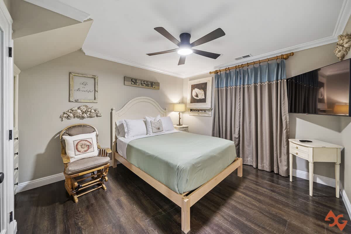 Inviting bedroom with a white queen-sized bed, dark wood flooring, a ceiling fan, and coastal-themed decor including a "SEASIDE" sign and shell artwork.