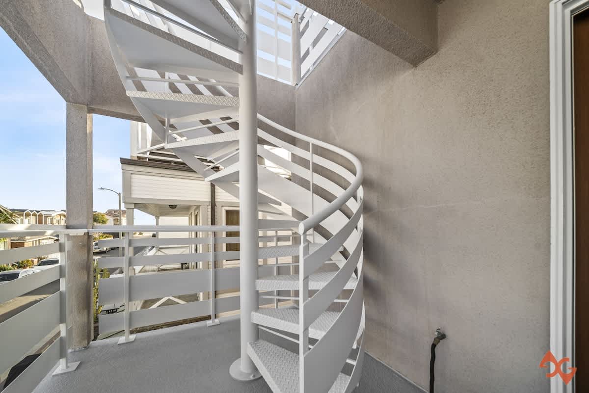 A white metal spiral staircase with open risers connects outdoor levels, set on a gray concrete surface with a white slatted railing. The stucco wall and a glimpse of the sky are visible.