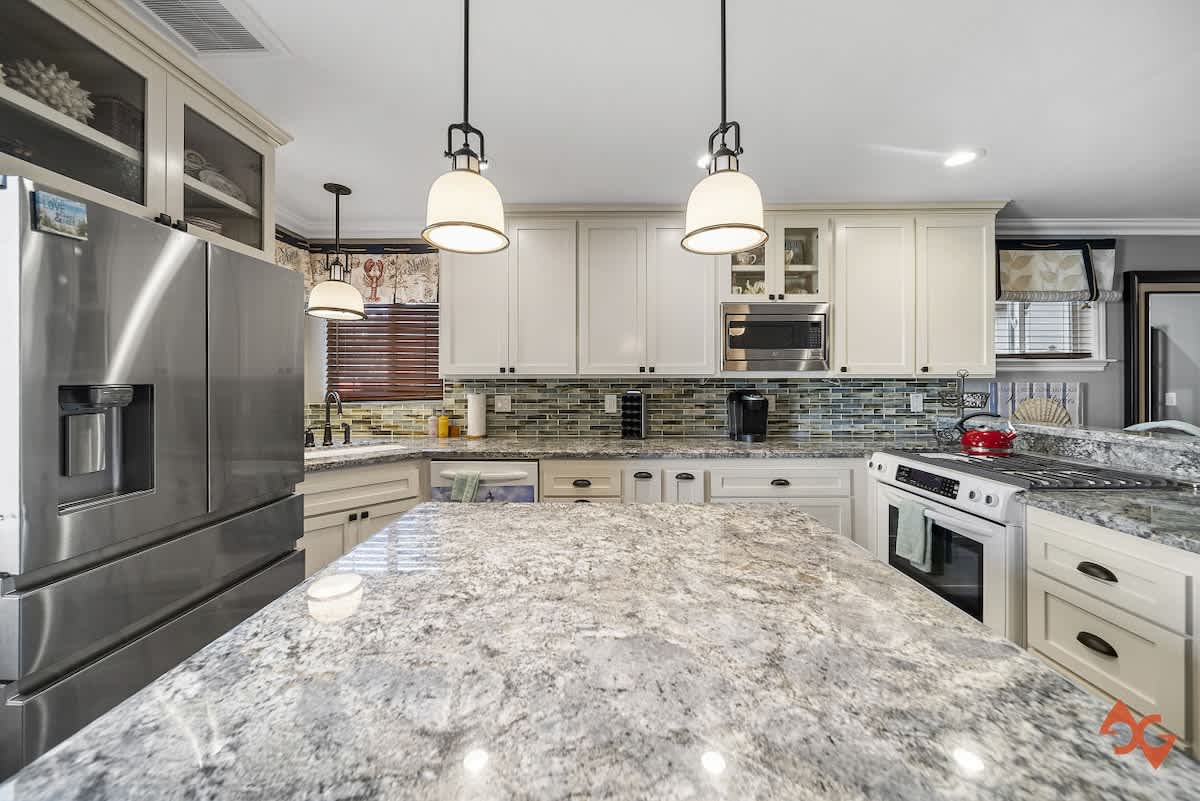 Modern kitchen with a large granite-topped island, stainless steel refrigerator, and white cabinets, featuring a green glass subway tile backsplash and pendant lighting.