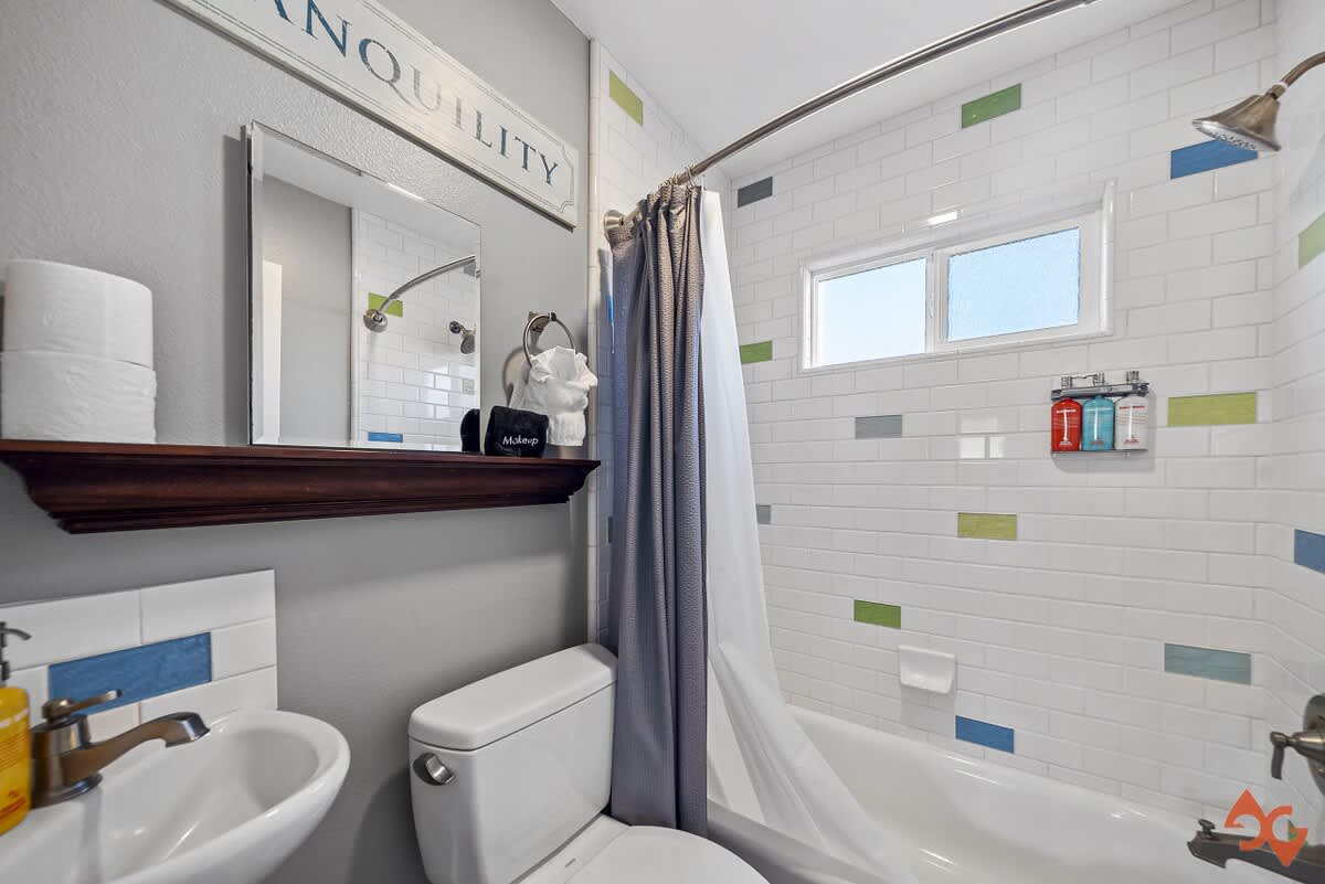 A bright bathroom with white subway tile walls featuring colorful accent tiles in blue, green, and gray, a white bathtub, and a dark wood floating shelf above the toilet.