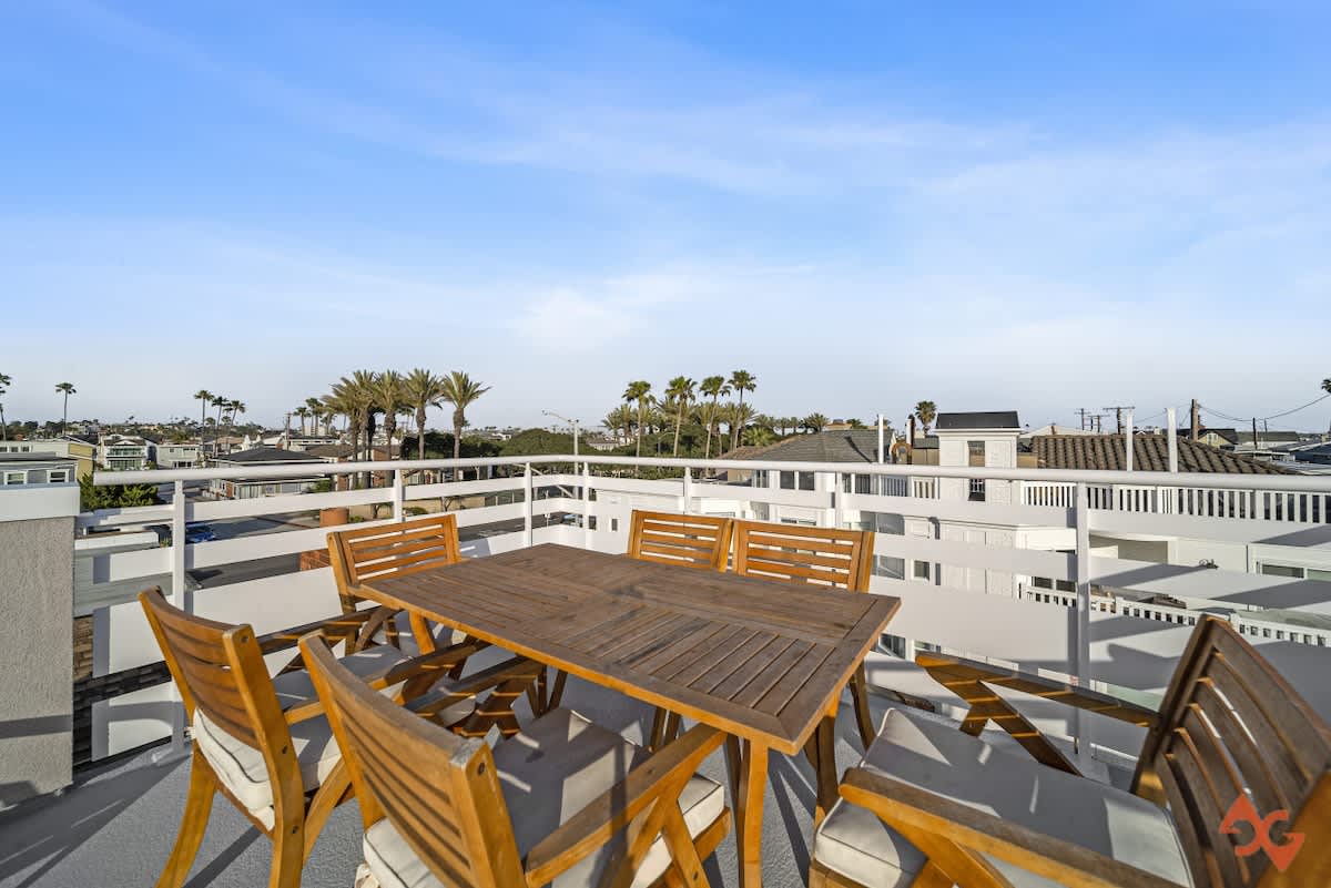 A wooden outdoor dining table with six matching chairs, featuring light cushions, is centered on the rooftop deck, with a white slatted railing and a view of distant rooftops and palm trees under a bright sky.