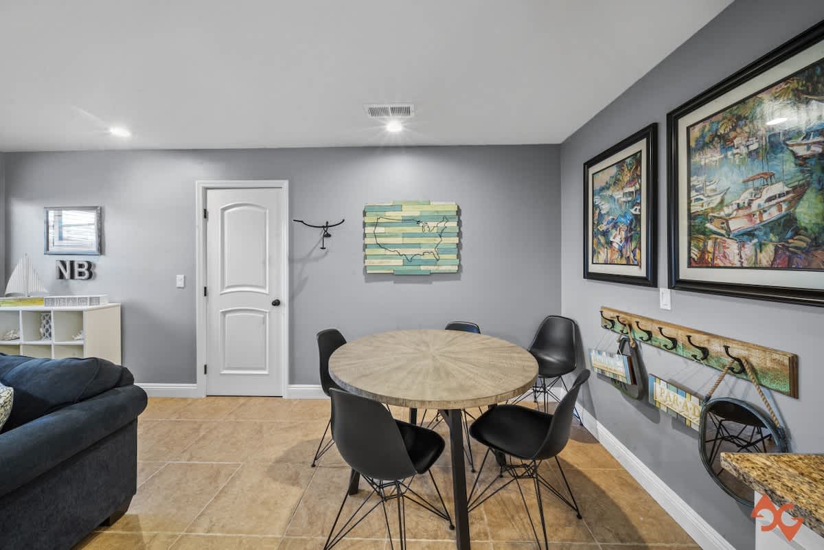Dining area with a round wooden table and black chairs, adjacent to a living space with a blue sofa, all on light tile flooring, featuring coastal-themed artwork.