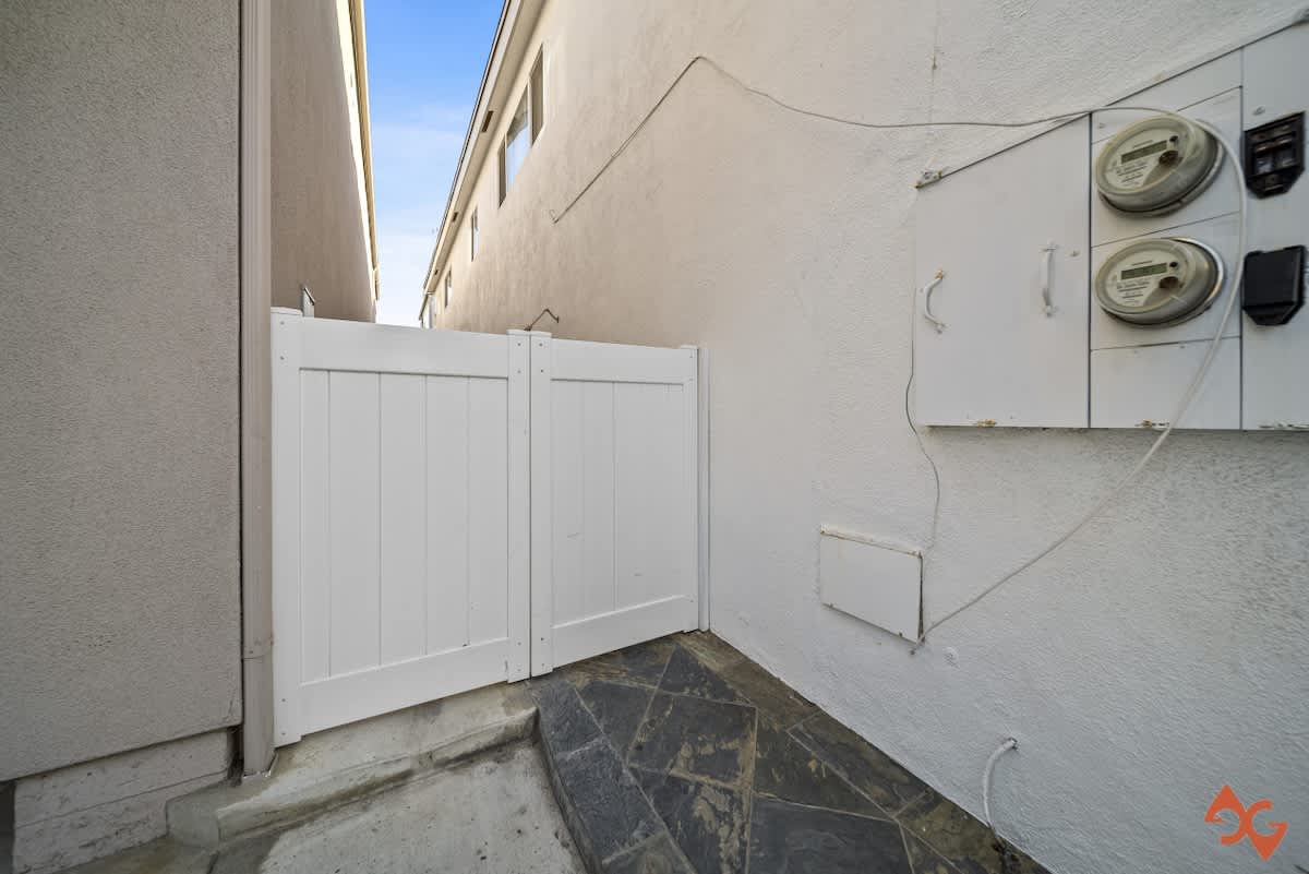 A white privacy gate stands at the end of a stone-tiled and concrete path, with utility meters and electrical boxes mounted on the adjacent white stucco wall.