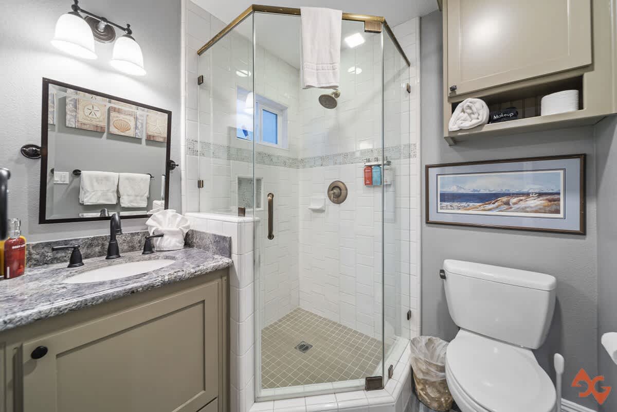 A modern bathroom featuring a glass-enclosed corner shower with white subway tiles and a small window. A vanity with a granite countertop and dark cabinet is on the left, and a toilet is on the right.