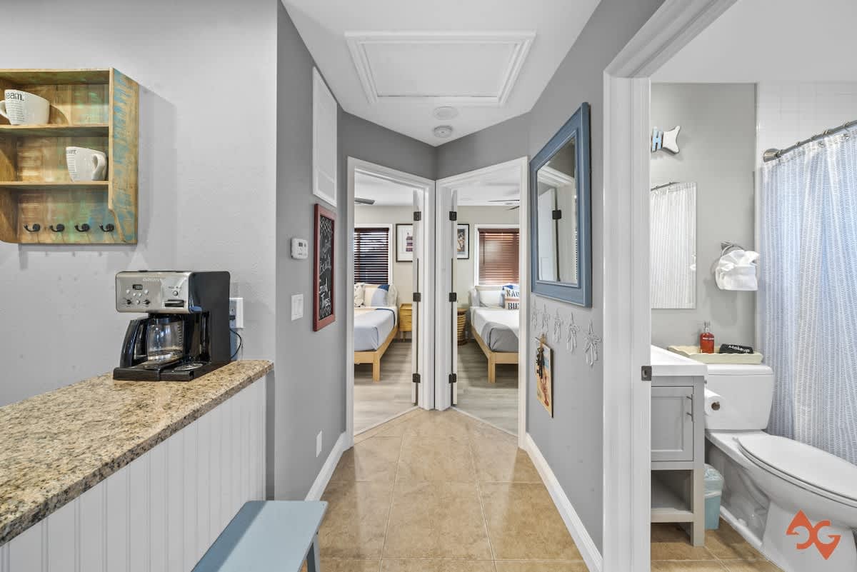 A hallway with light gray walls and tile flooring, featuring a coffee bar area with granite countertops and a glimpse into a bathroom with a toilet and shower curtain.