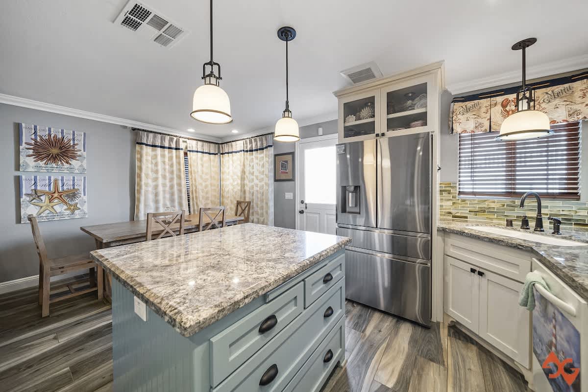 Well-appointed kitchen featuring a light blue island with granite top, stainless steel refrigerator, and white cabinetry, with a dining area visible in the background.
