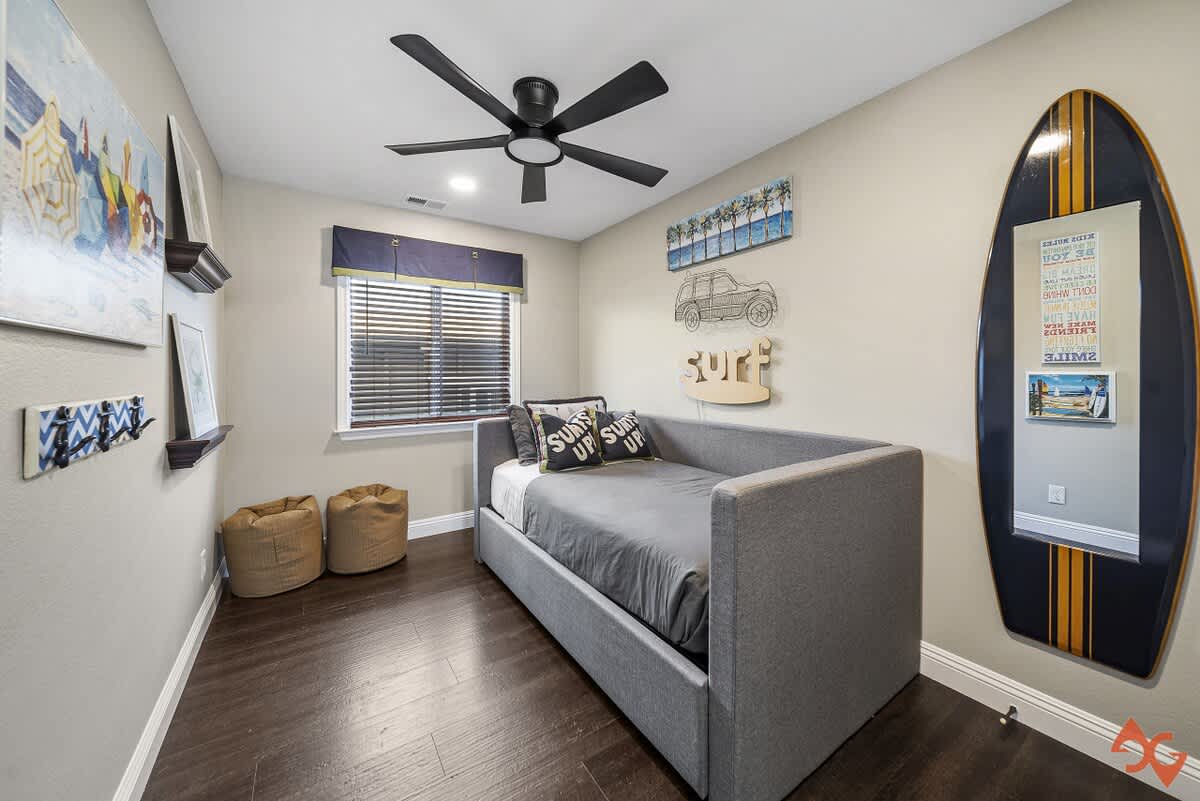 Kids' bedroom featuring a gray daybed with 'SURFS UP!' pillows, dark wood floors, a large surfboard mirror, and surf-themed wall art, including a metal car and 'surf' sign.