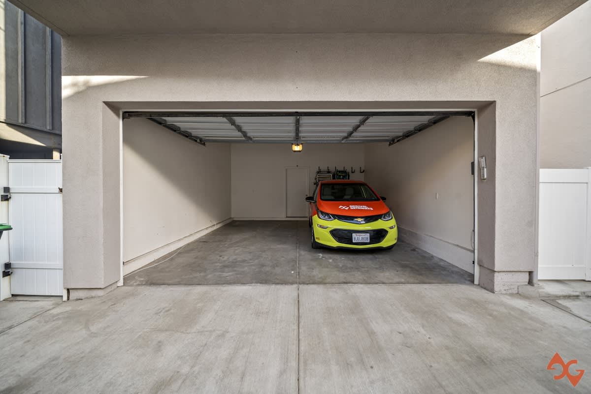 A wide view of an open garage entrance with a concrete driveway leading in, showcasing a vibrant orange and yellow car parked within the clean, white-walled interior.