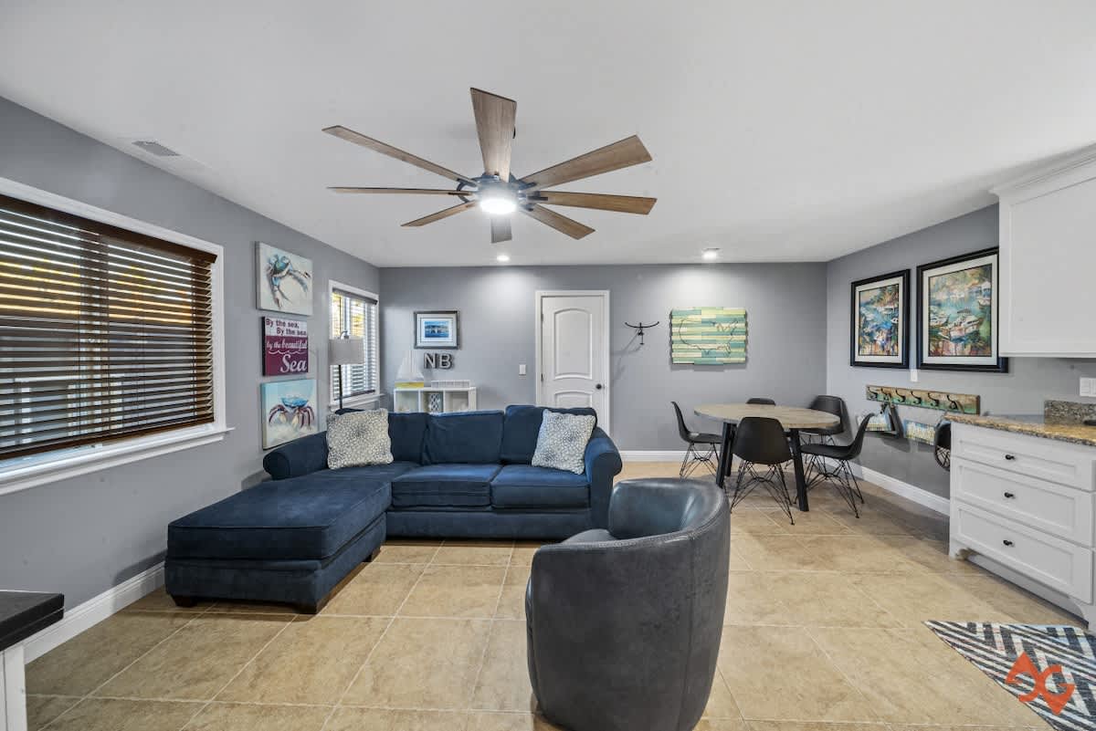 A modern living room features a large navy blue sectional sofa, a gray armchair, and a round dining table with black chairs, all set on light brown tiled floors under a large ceiling fan. Gray walls are adorned with coastal-themed art.