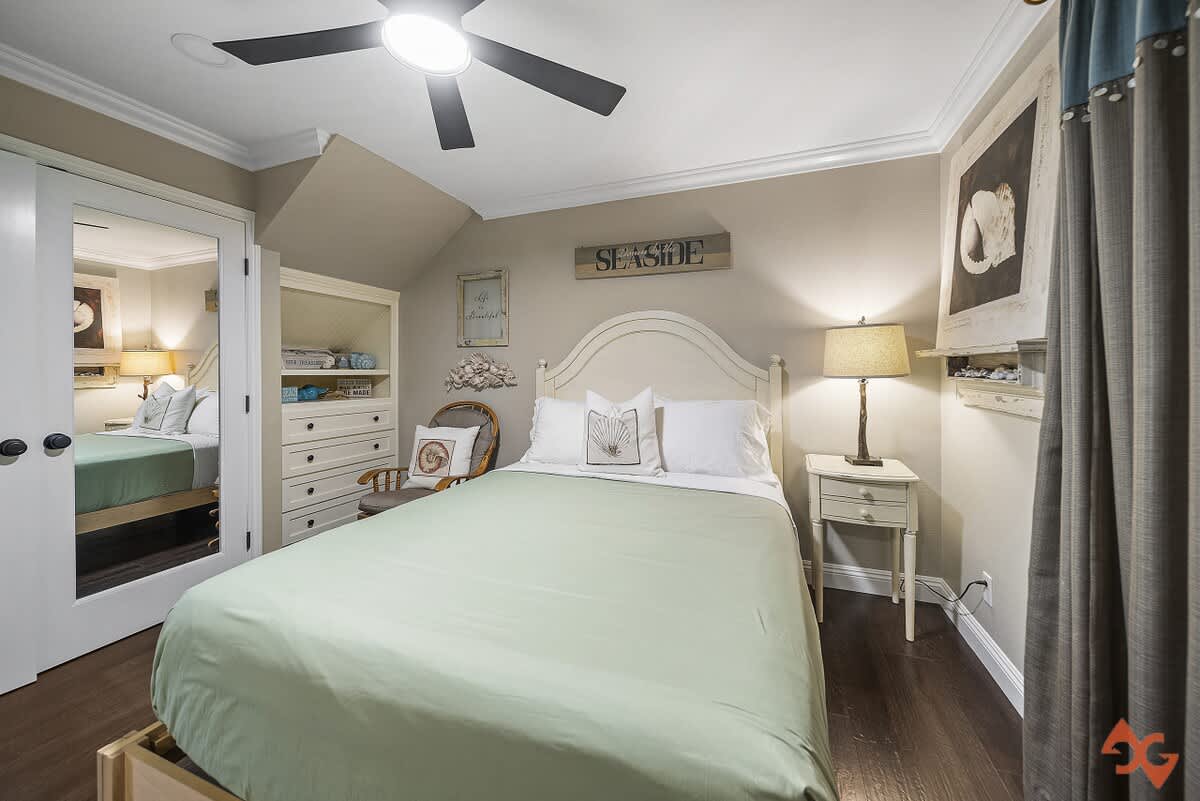 Cozy bedroom with a white headboard bed topped with a light green duvet, dark wood floors, a white nightstand with a lamp, and a white dresser with open shelving, featuring coastal-themed decor.