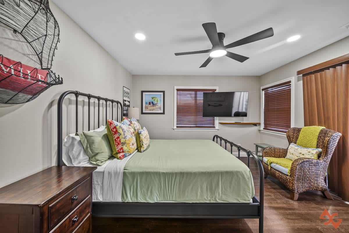 A bedroom with a black metal bed frame, dark wood nightstand, and a wicker armchair, featuring a wall-mounted TV and a window with wooden blinds, all under a black ceiling fan.