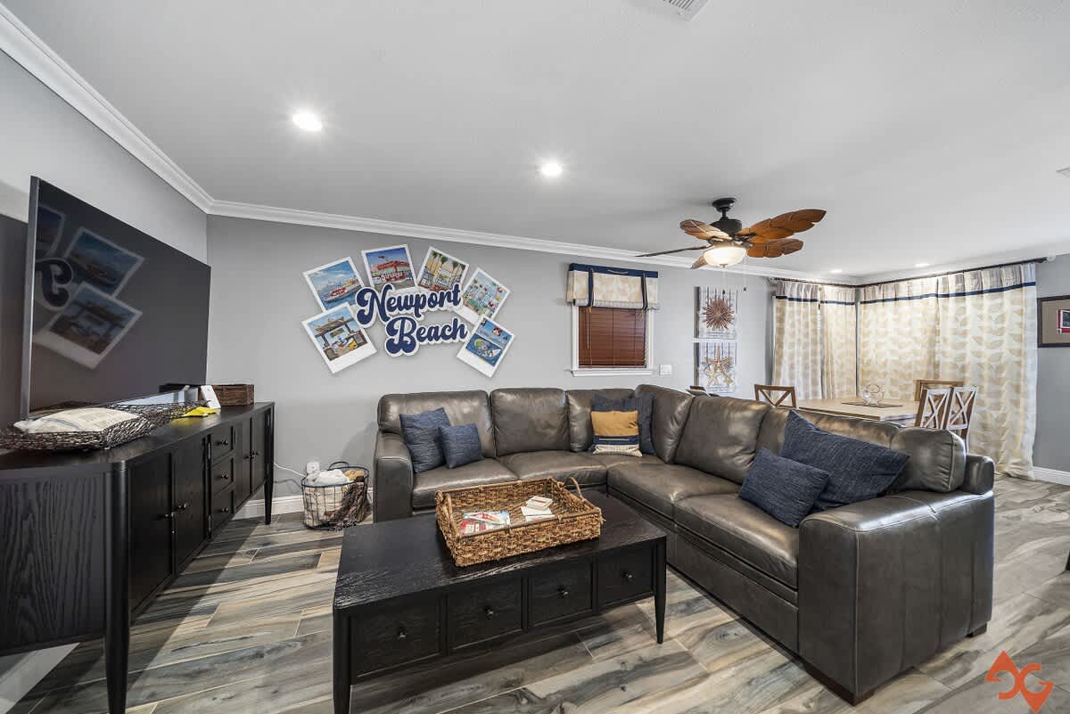 Spacious living room with a large dark gray leather sectional sofa, a black coffee table, and a wall-mounted TV, featuring wood-look flooring and coastal-themed wall art.