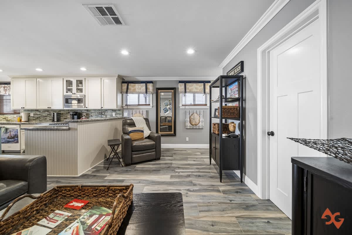 An open-concept living area with wood-look tile flooring, a gray sectional sofa, and a view into the modern kitchen with white cabinets and a large island. A leather recliner and a black shelving unit are also visible.