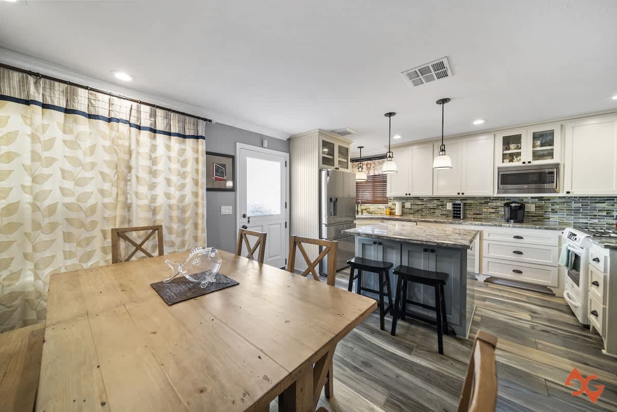 A bright kitchen and dining area with wood-look tile flooring, white cabinets, a large island with bar stools, and a wooden dining table with chairs. A decorative fish centerpiece is on the table.