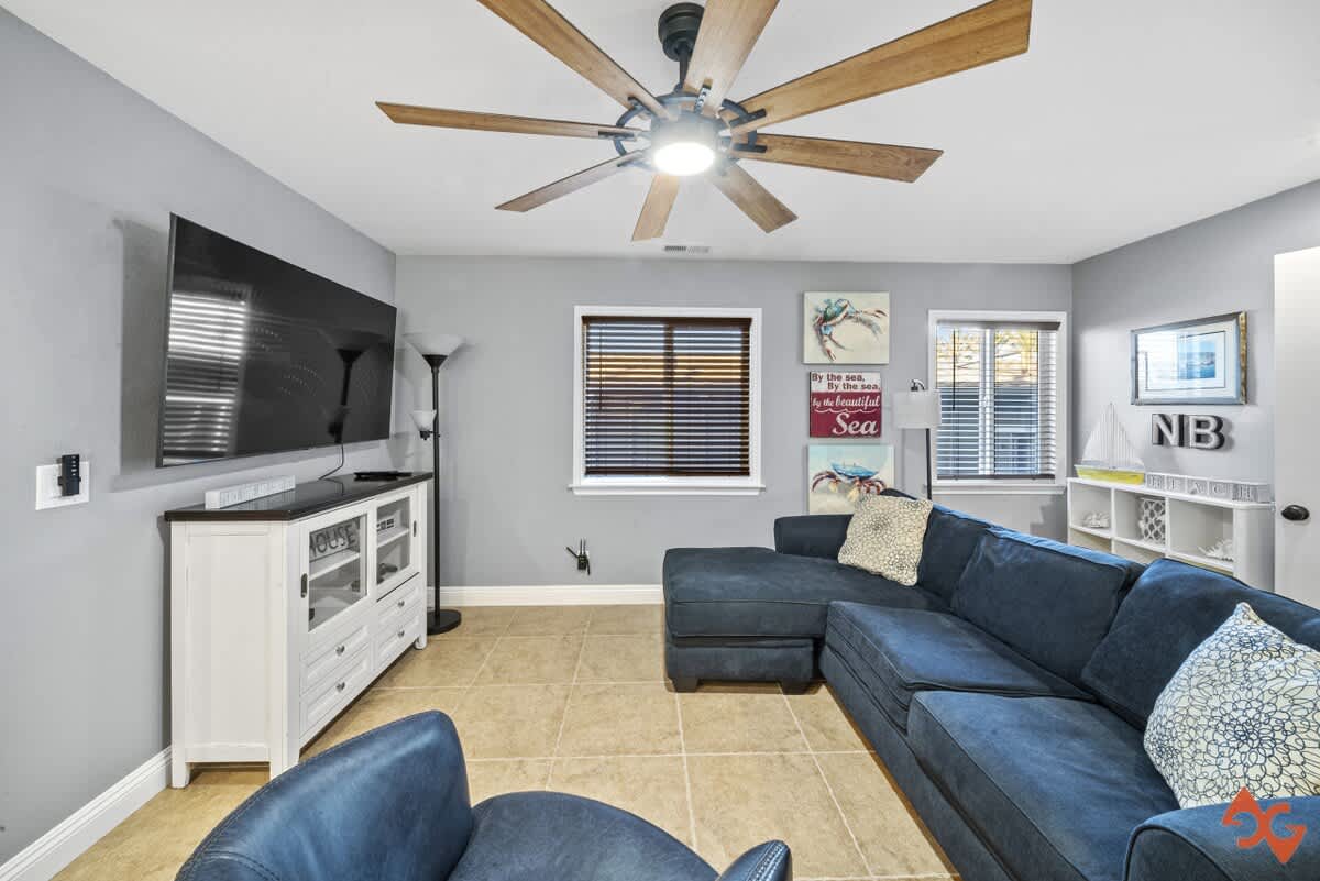 A comfortable living room with light gray walls, a large blue sectional sofa, a white media console with a wall-mounted TV, and a large ceiling fan, all on light-colored tile flooring.