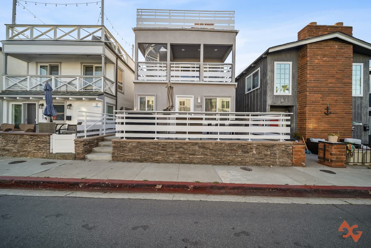 Three-story vacation rental house with a light gray facade and white railings, featuring multiple balconies and a ground-level patio, situated directly on a street.