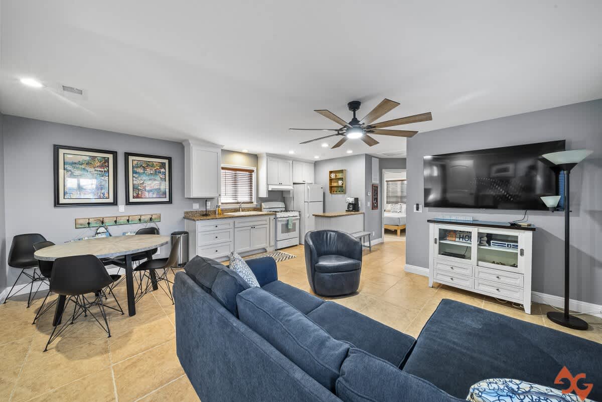 Open-concept living area with a blue sectional sofa, a round dining table with black chairs, and a white kitchen with granite countertops, all on light tile flooring.