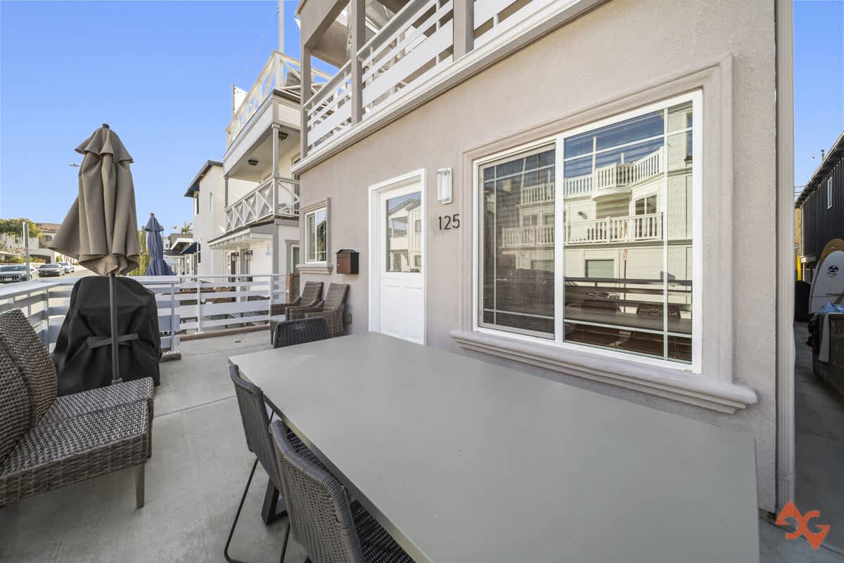 The exterior of a light-colored building with white trim and multiple windows, featuring a ground-level patio with an outdoor dining table and chairs. The address "125" is visible next to a white door.