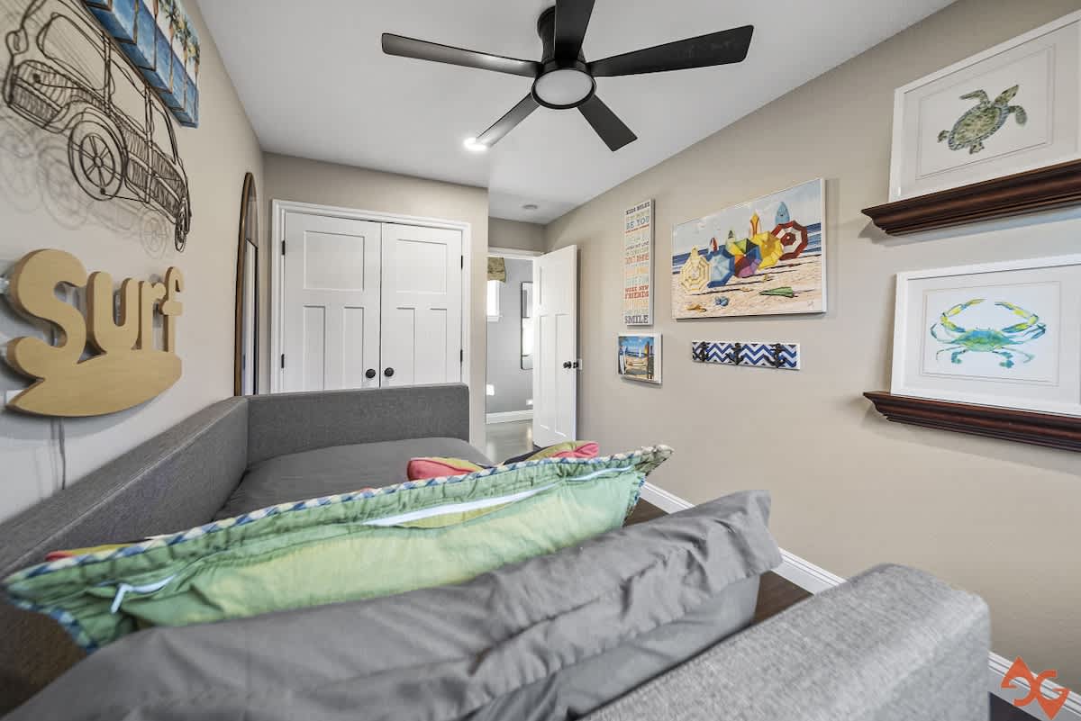 A cozy bedroom featuring a gray daybed with colorful pillows, a black ceiling fan, and beach-themed wall art including a "Surf" sign and a vintage car illustration.