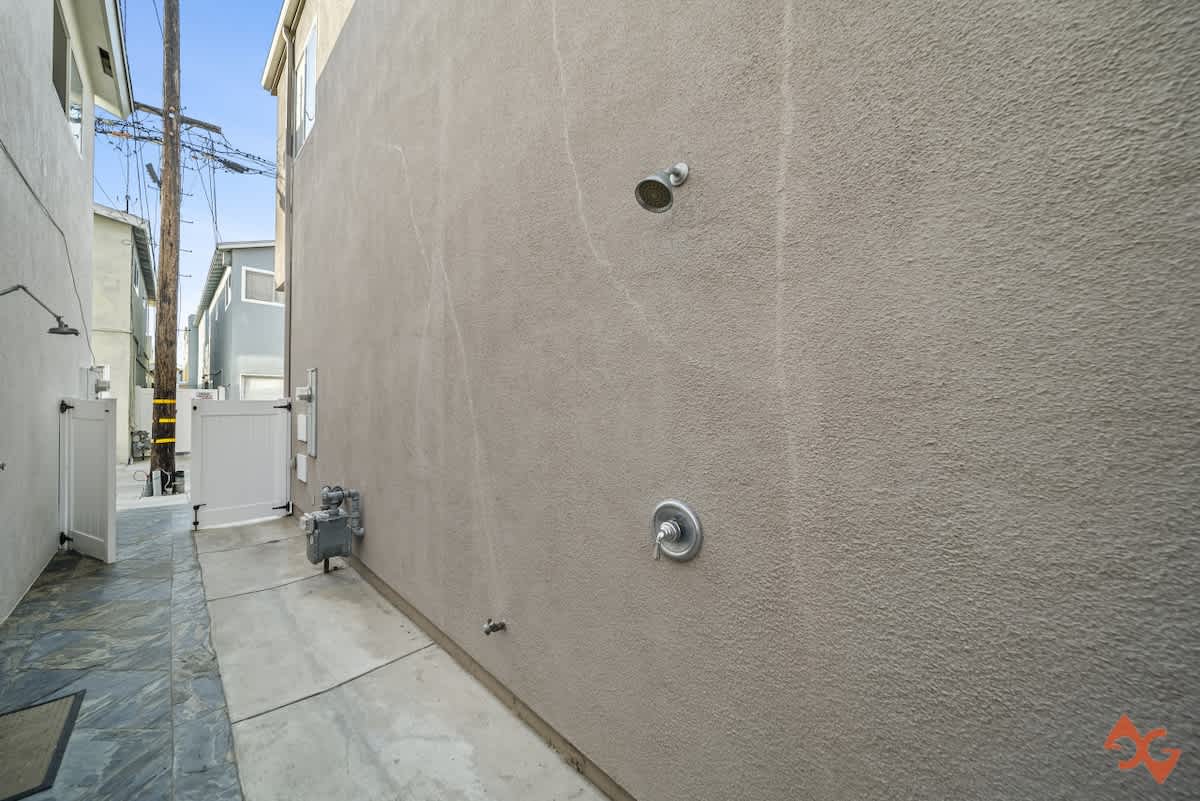 A narrow concrete path runs alongside a textured stucco wall with an outdoor shower, leading towards a white gate and utility poles in the distance under a clear sky.