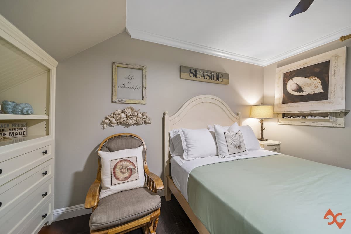 A bright bedroom with dark wood floors and a light green bedspread on a white bed frame. A white dresser with open shelving is on the left, and a nightstand with a lamp is on the right, adorned with coastal decor.