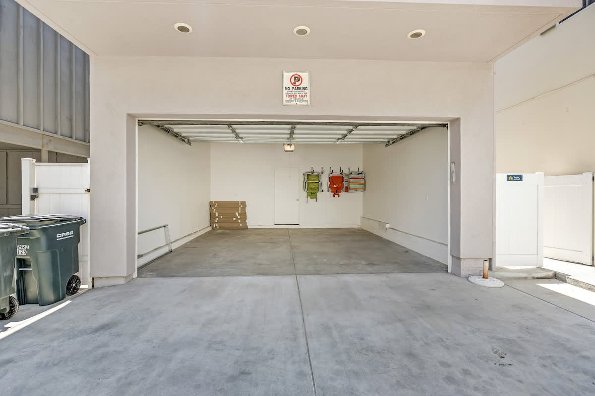 A spacious, empty two-car garage with a concrete floor and white walls, featuring an open garage door and a 'No Parking' sign. Several colorful beach chairs are hung on the back wall.