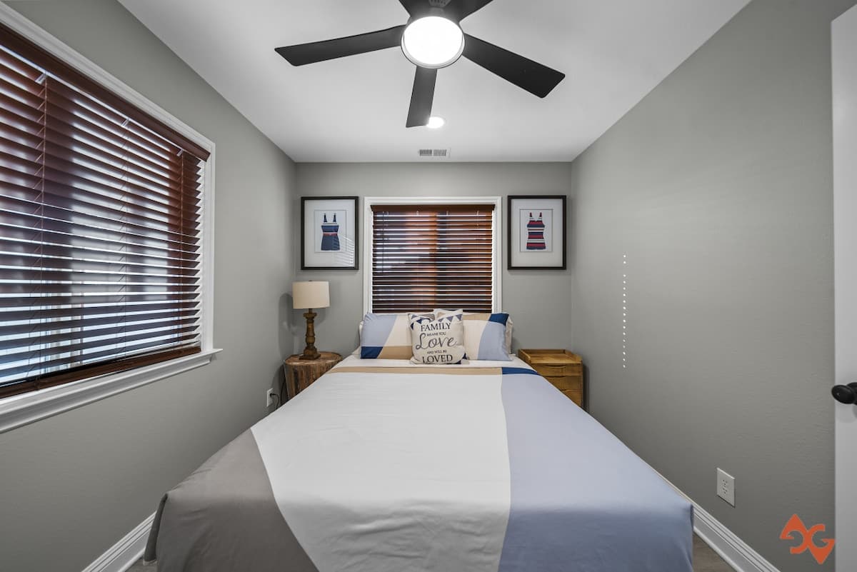 A narrow view of bedroom-2, featuring a queen bed with blue and white bedding, a wooden nightstand with a lamp, and two windows with dark wood blinds. Framed swimsuit art hangs above the bed.