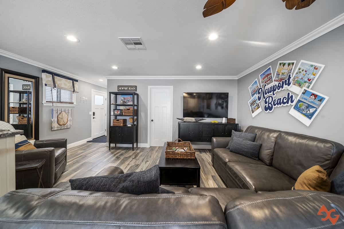Spacious living room featuring a large dark gray leather sectional sofa, a black coffee table with a woven basket, and a wall-mounted TV, all set on light wood-look flooring with coastal-themed wall art.