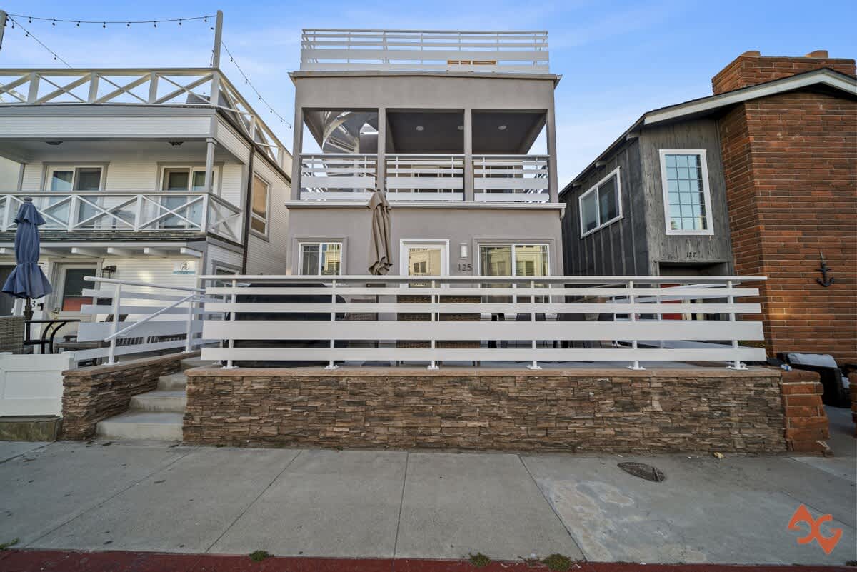 The front exterior of a three-story house with a light gray stucco finish, featuring a white horizontal slatted fence on a stone base, and a top-level balcony with a spiral staircase visible on the left.