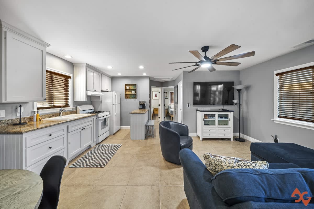 An open-concept living space featuring a modern kitchen with white shaker cabinets and granite countertops, flowing into a living area with a large blue sectional sofa and a ceiling fan.